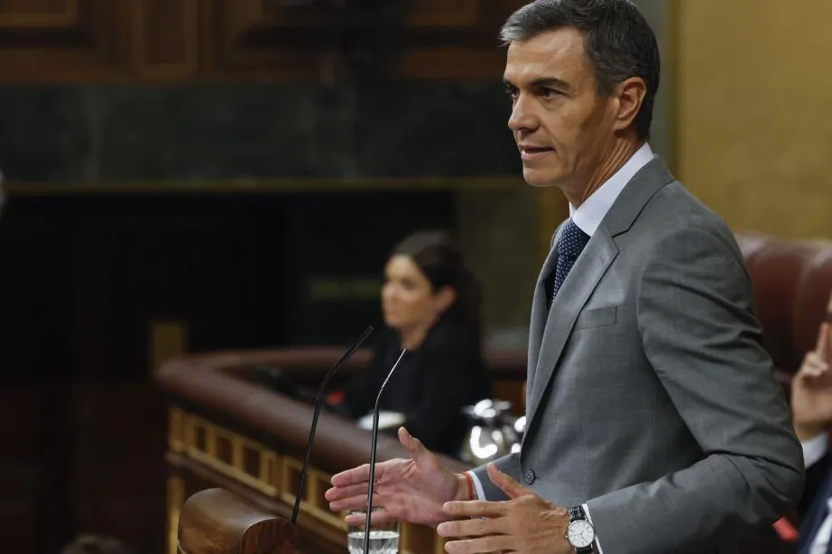 Fotografía de Pedro Sánchez interviniendo desde la tribuna del Congreso de los Diputados, con una mujer al fondo y micrófonos en primer plano.