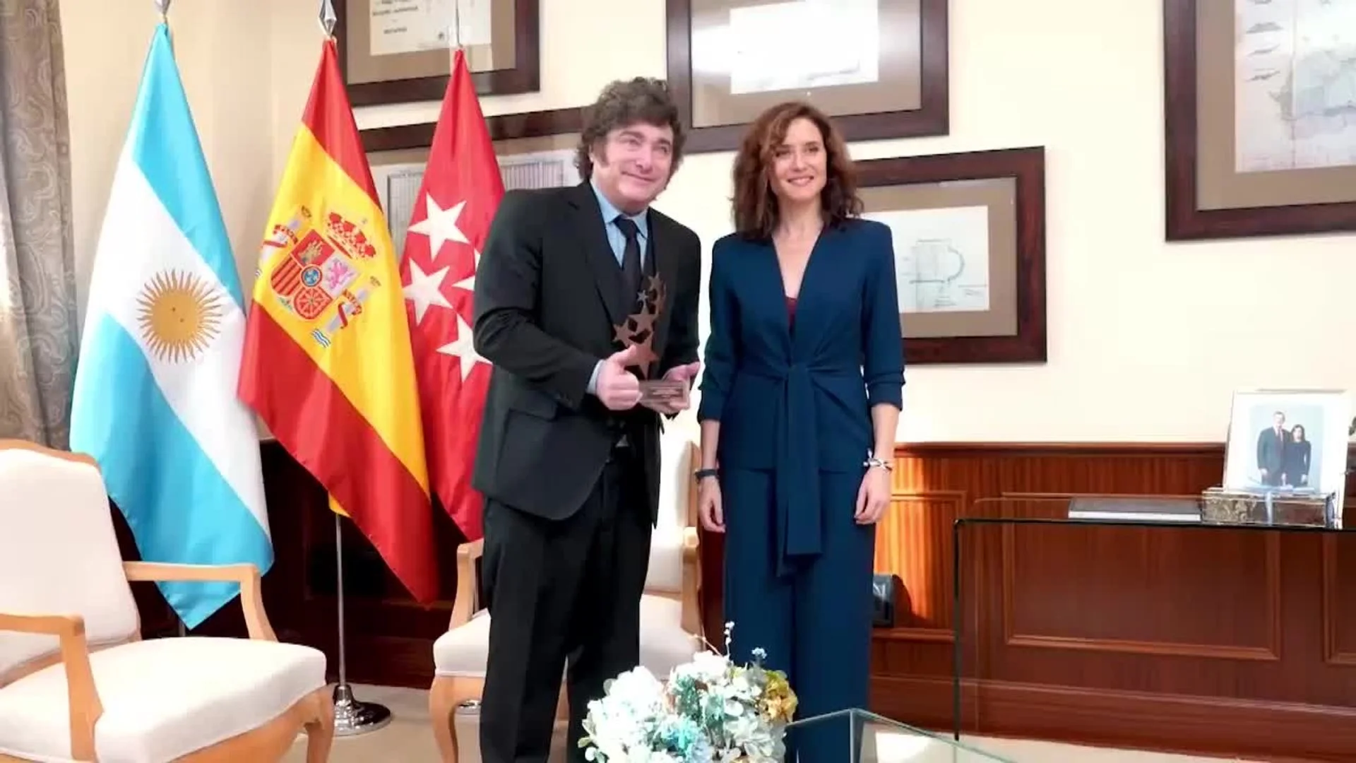 Javier Milei y Isabel Díaz Ayuso posan juntos sonrientes en un despacho institucional con banderas de Argentina y España de fondo.