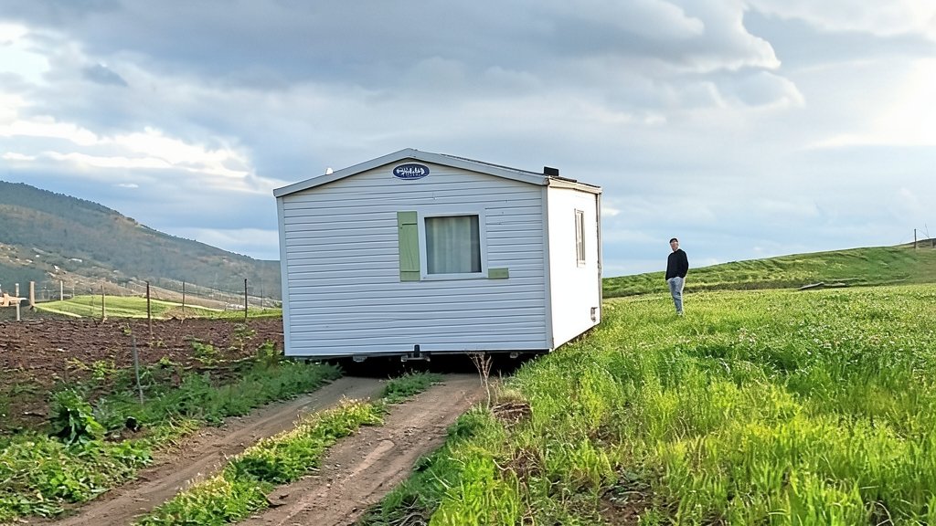 Fotografía que muestra una casa móvil blanca instalada en un terreno rural montañoso, con una persona de pie al lado y un camino de tierra.