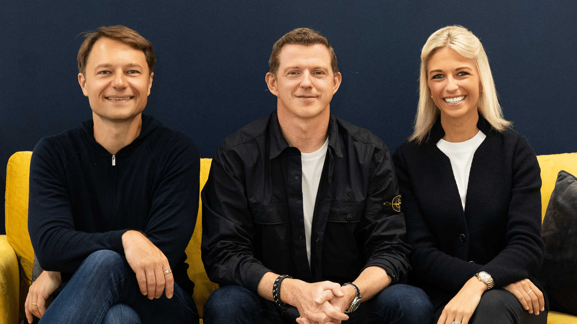 Fotografía editorial de tres cofundadores y socios de DIG Ventures (Rytis Vitkauskas, Ross Mason y Melissa Klinger) sentados en un sofá amarillo frente a una pared azul.