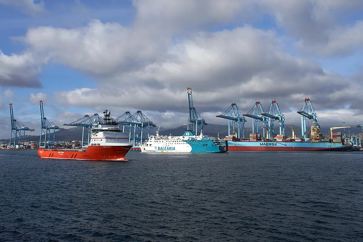 Vista panorámica del puerto de Algeciras con barcos, incluyendo un buque rojo en primer plano, un ferry de Balearia y un portacontenedores de Maersk atracados junto a grandes grúas pórtico bajo un cielo nublado.