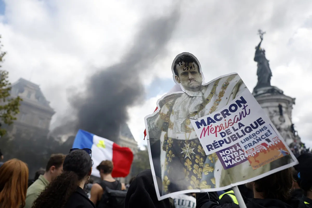 Protesta en París con manifestantes portando un cartel satírico de Emmanuel Macron vestido de monarca, acompañado de una bandera francesa y una columna de humo de fondo.