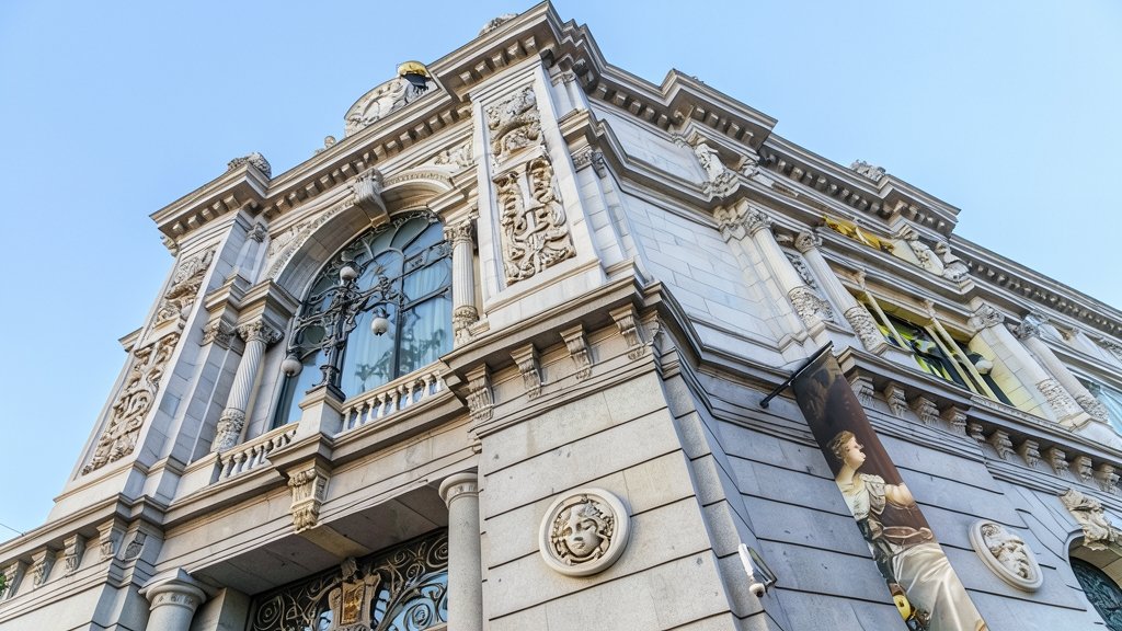 Fachada clásica del edificio del Banco de España con elementos arquitectónicos ornamentales, grandes ventanales y un cartel publicitario vertical.