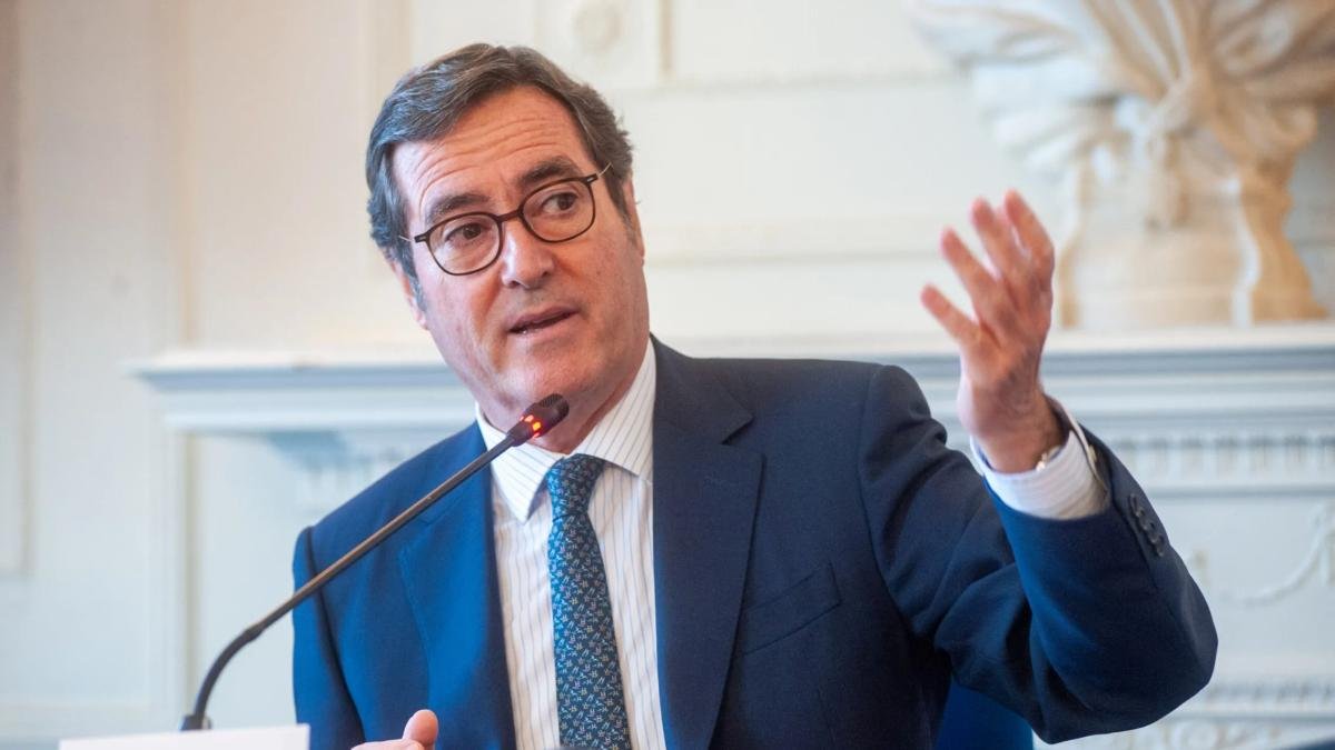 Fotografía editorial de Antonio Garamendi, presidente de la CEOE, interviniendo en un foro con traje oscuro, corbata estampada y gafas, gesticulando con la mano izquierda y con un micrófono de mesa delante.