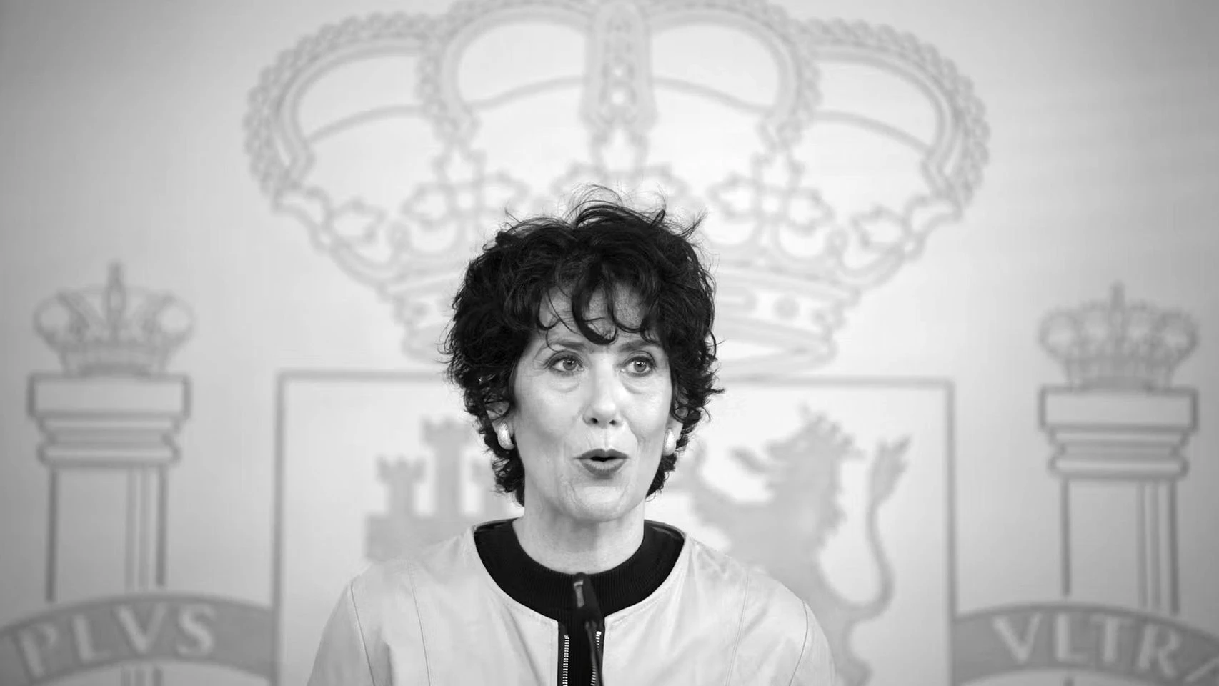 Fotografía en blanco y negro de una mujer hablando en una rueda de prensa institucional con el escudo de España de fondo.