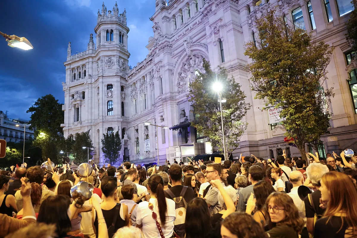 Fotografía nocturna de una multitud manifestándose frente a un edificio institucional en Madrid con pancartas y cacerolas.
