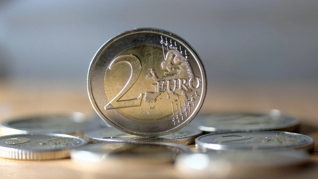 Moneda de 2 euros en primer plano vertical apoyada sobre otras monedas dispersas en una superficie de madera con fondo neutro.