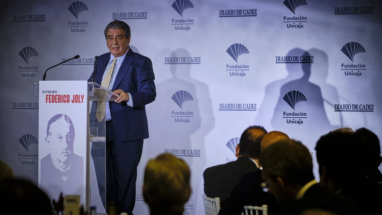 Fotografía editorial de Augusto Delkáder interviniendo en un atril con el logotipo del Diario de Cádiz y la Fundación Unicaja durante un evento conmemorativo.