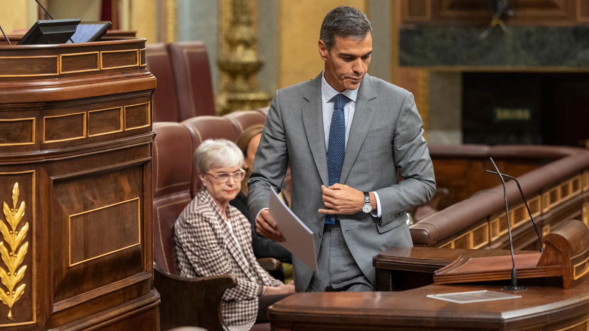 Pedro Sánchez con traje gris y corbata azul en el Congreso de los Diputados sosteniendo unos documentos.