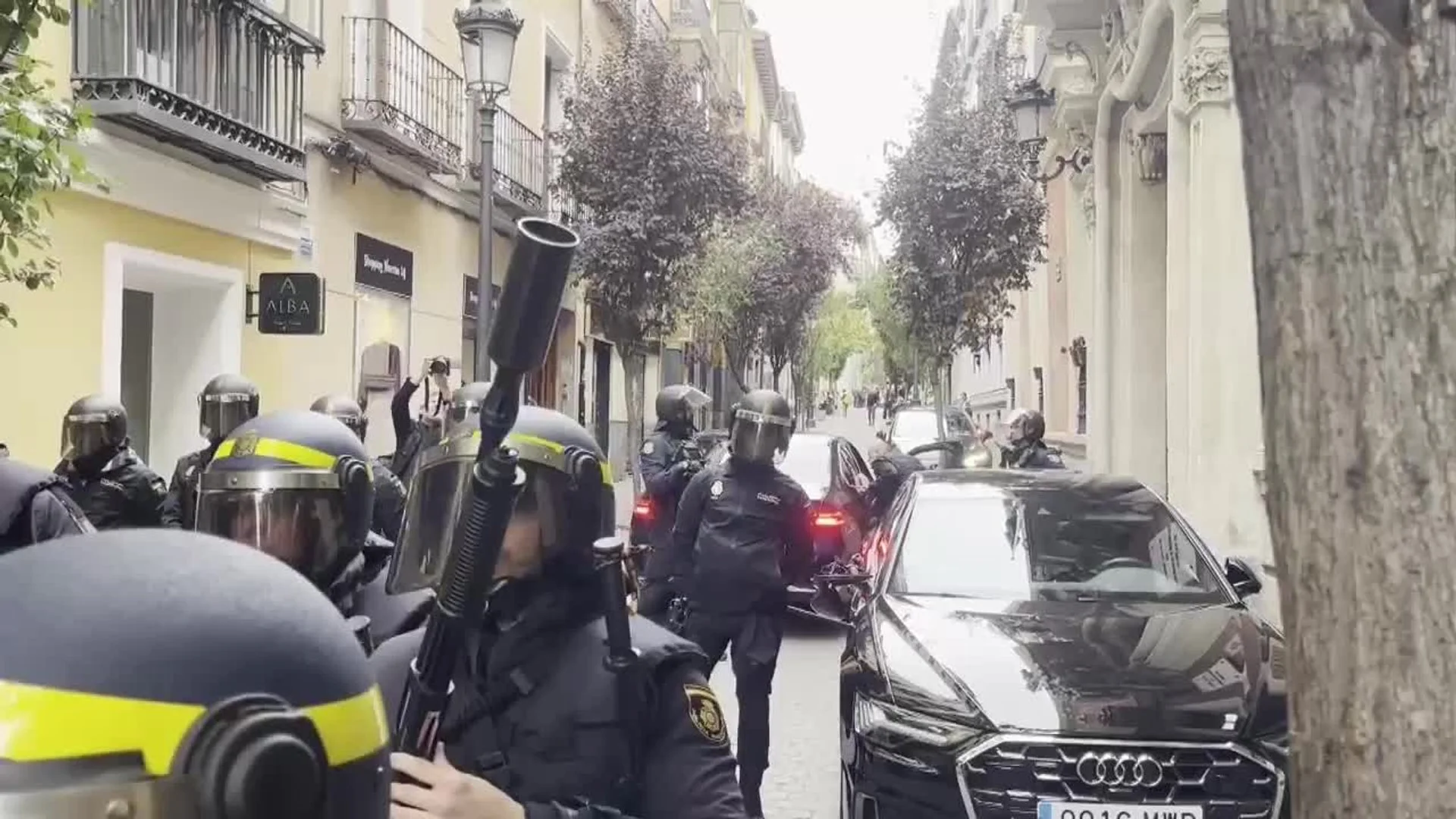 Fuerzas policiales antidisturbios con cascos y equipamiento táctico desplegados en una calle estrecha de Madrid junto a vehículos oficiales.