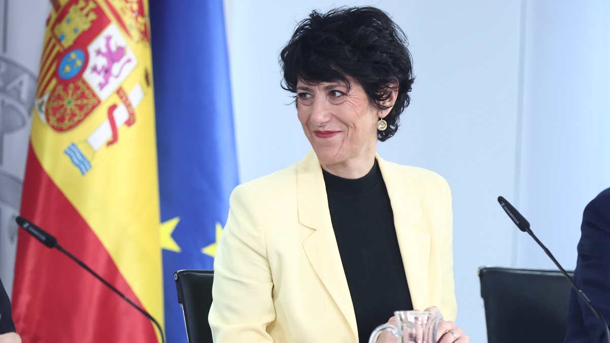 La ministra Elma Saiz sonriendo durante una comparecencia con banderas institucionales de fondo.