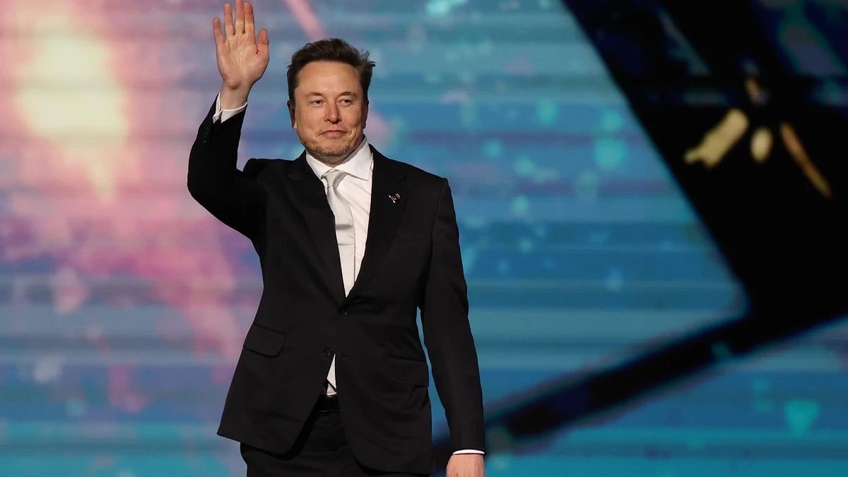 Elon Musk saludando con la mano derecha en un escenario con un traje oscuro y corbata plateada, con pantallas de fondo.