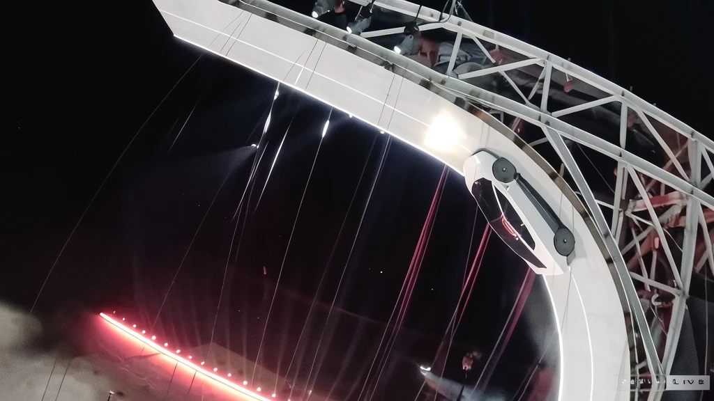Un coche blanco subiendo por una rampa curva iluminada de noche con luces rojas en el suelo y una estructura metálica al fondo.