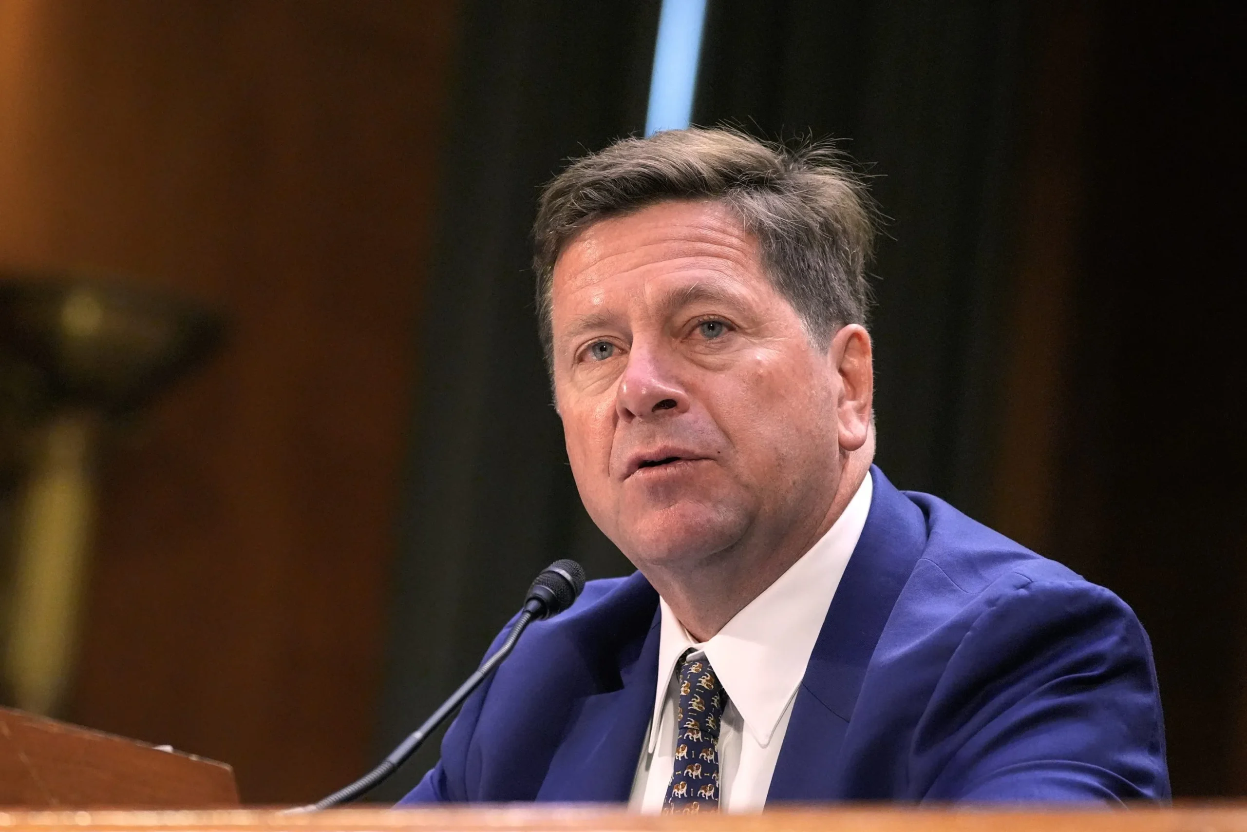 Fotografía editorial de Jay Clayton vistiendo traje azul y corbata con patrones sentado ante un micrófono en una sala de audiencias.