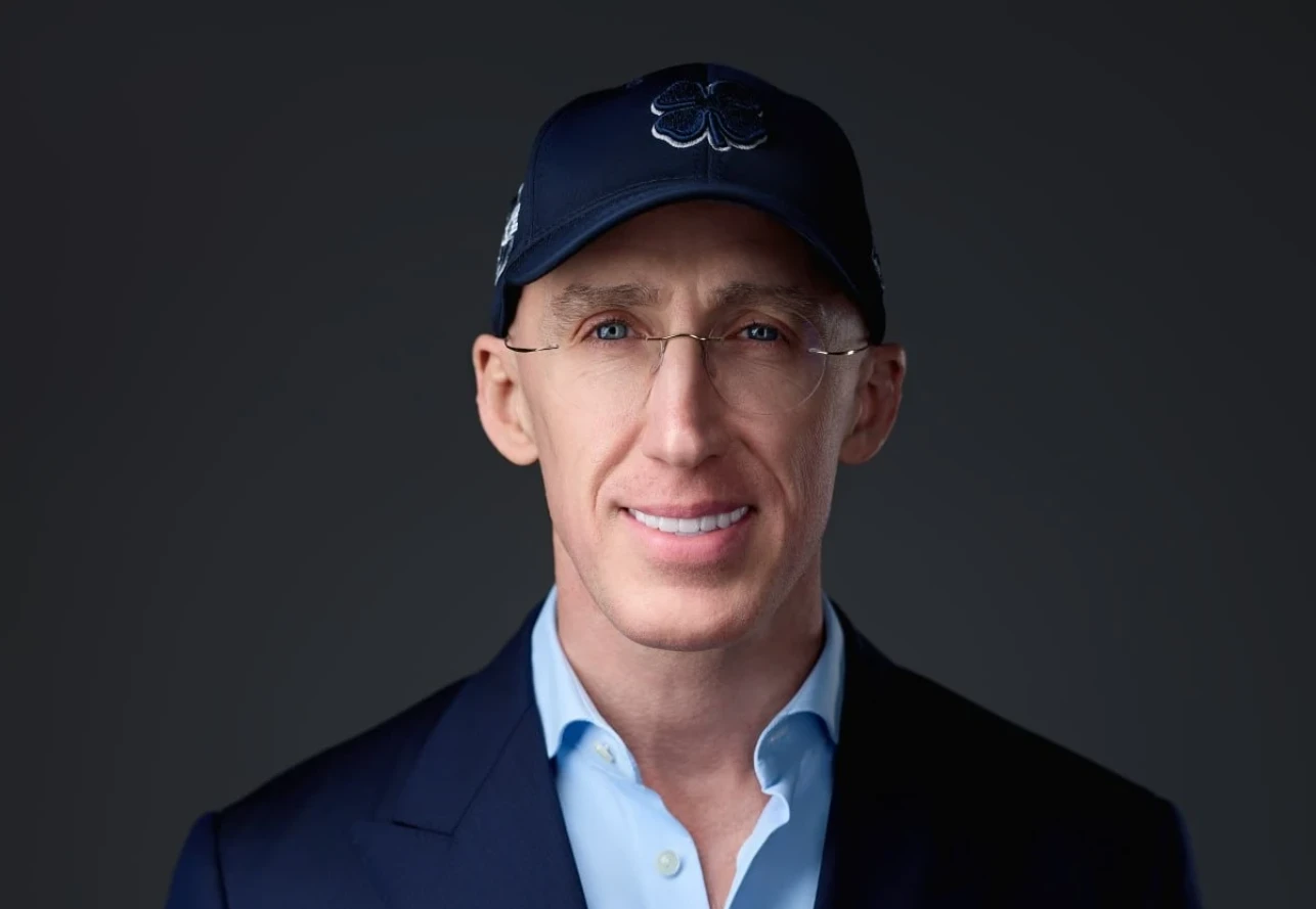 Retrato de estudio de Uri Poliavich con gorra oscura con un trébol bordado, gafas minimalistas, camisa azul claro y chaqueta oscura sobre fondo gris neutro.